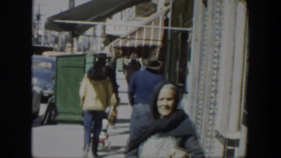1951 Texas town sidewalk frame with a headscarfed woman facing camera, pedestrians beyond her, storefronts, awnings, and a curbside car.