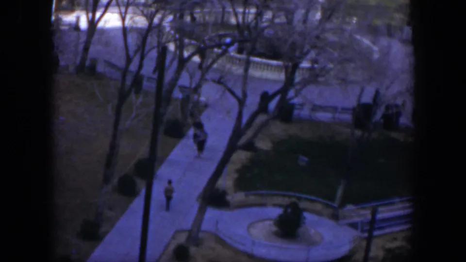 High-angle 1951 Texas home-movie frame showing pedestrians on a shaded courtyard walkway beside white railings and bare trees.