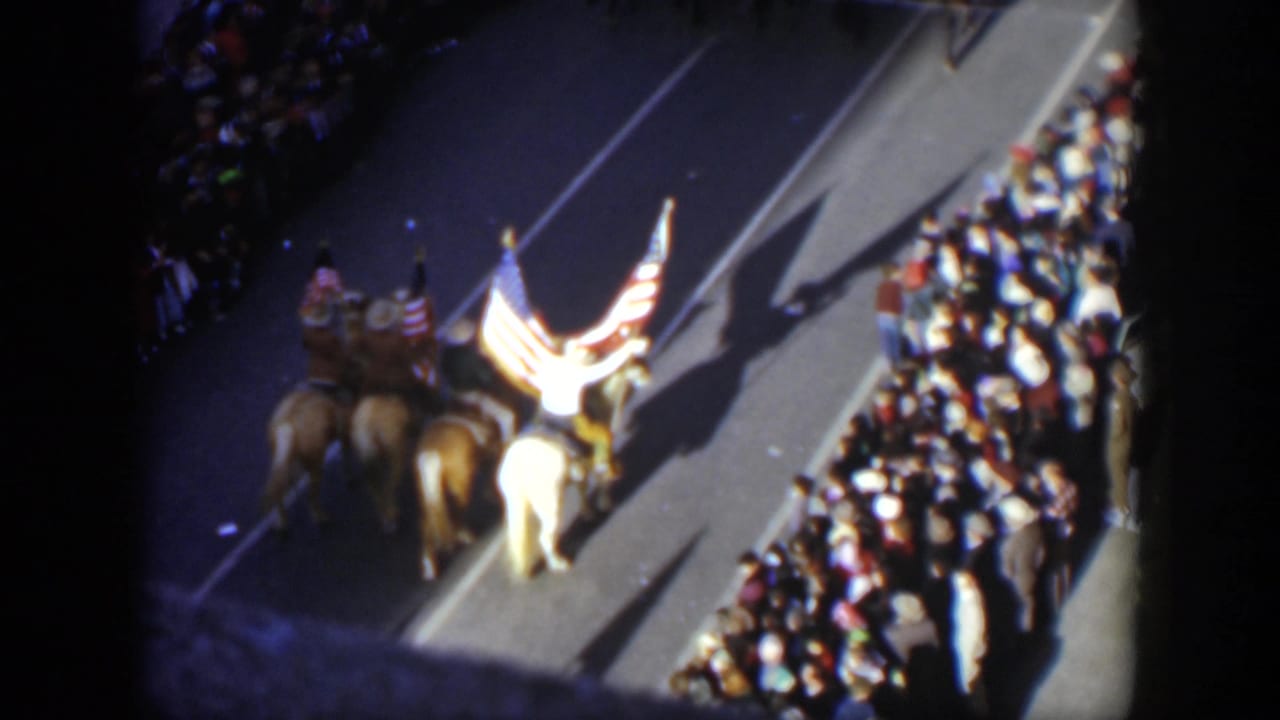 Mounted riders carrying United States flags pass a crowd-lined street.