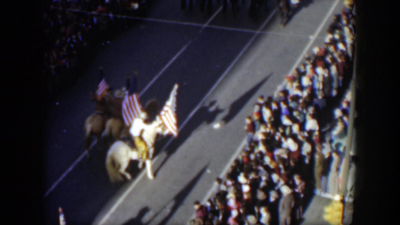 Riders on horses carry large United States flags beside a packed sidewalk.