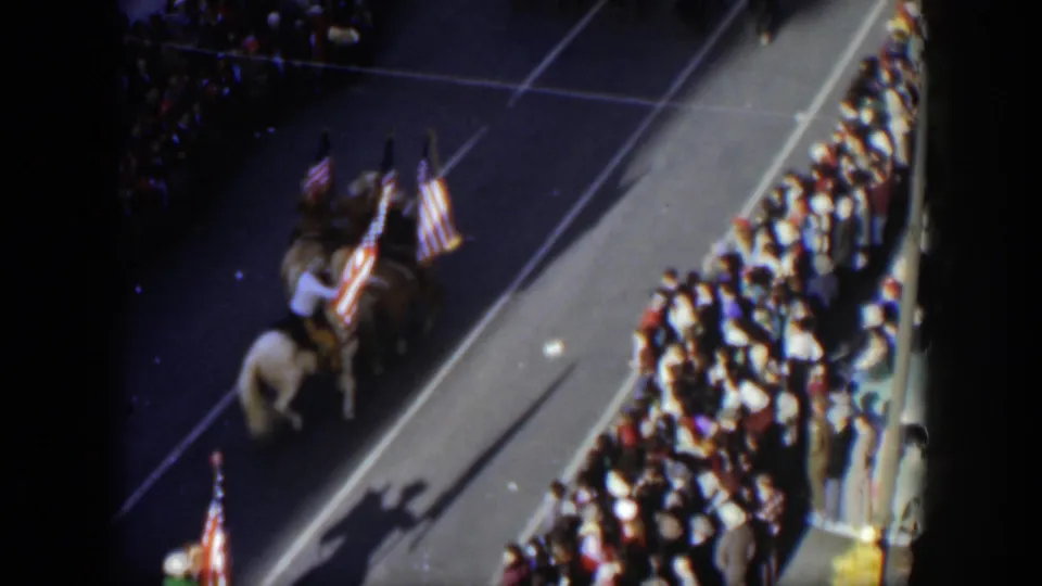 A high-angle home-movie frame shows horseback riders with United States flags moving past a crowded Texas parade route.