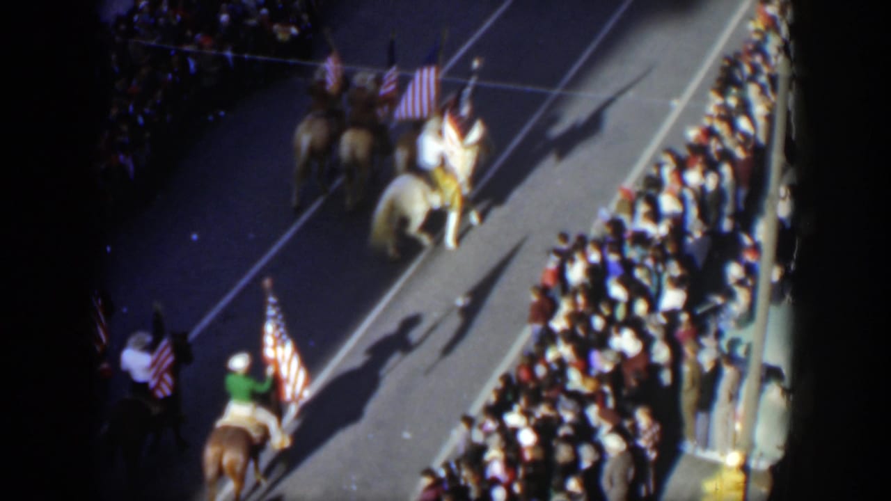 High-angle 1951 Texas 8mm frame of mounted parade riders with American flags moving past dense sidewalk spectators.
