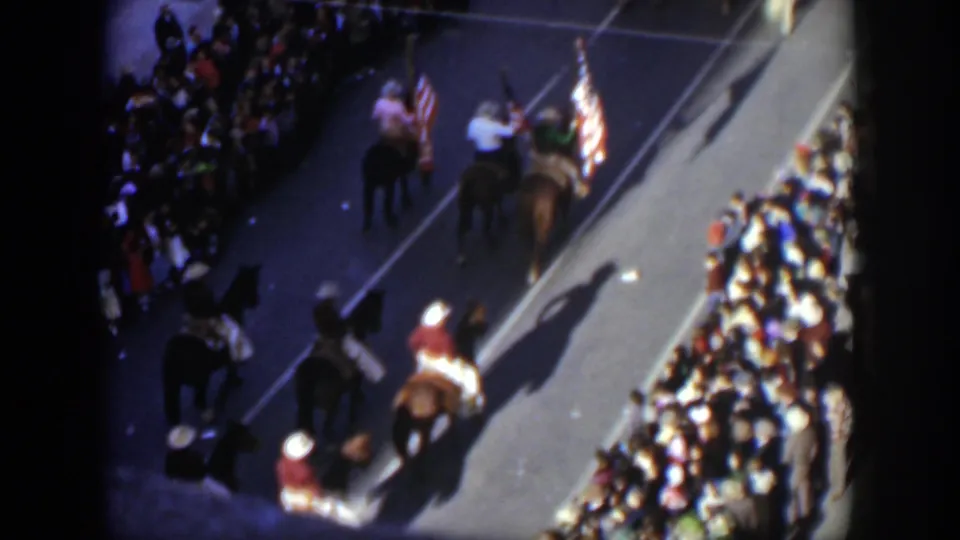 High-angle 1951 Texas parade frame with riders on horseback, American flags, strong shadows, and spectators lining the street.
