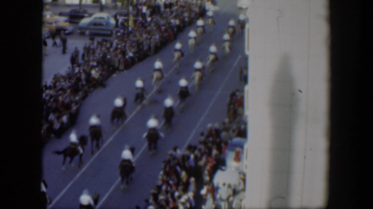 1951 Texas home-movie frame showing riders on horses moving in formation past dense sidewalk crowds and parked cars.