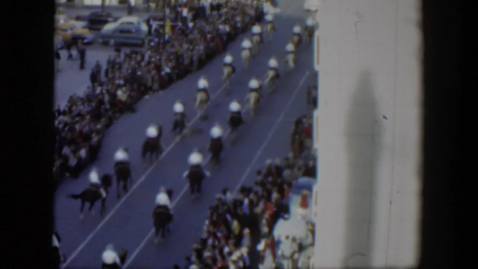 1951 Texas home-movie frame showing riders on horses moving in formation past dense sidewalk crowds and parked cars.