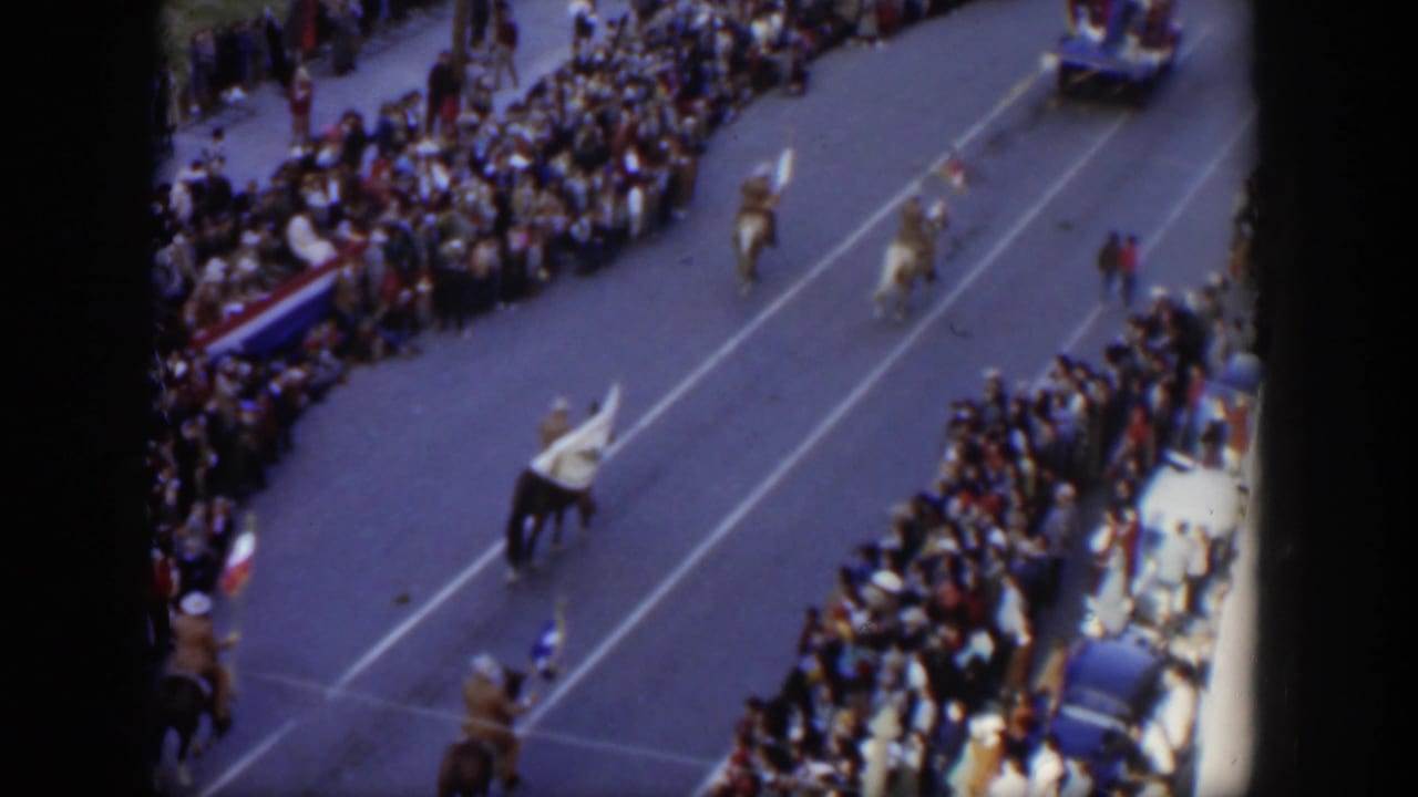 Mounted riders carrying flags move along a crowded city parade route viewed from above.