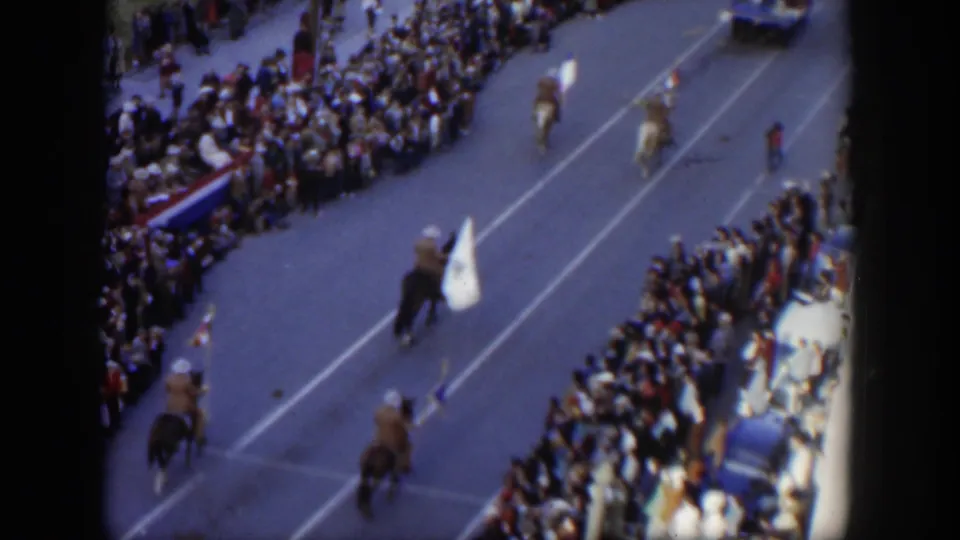 High-angle 1951 Texas parade view with riders on horseback carrying flags past packed sidewalks and a visible reviewing area.