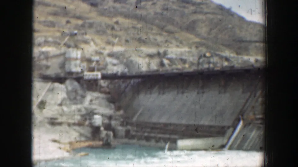 1937 California 8mm frame showing a concrete dam section with steel truss bridgework, service structures, dry hills, and reservoir water.