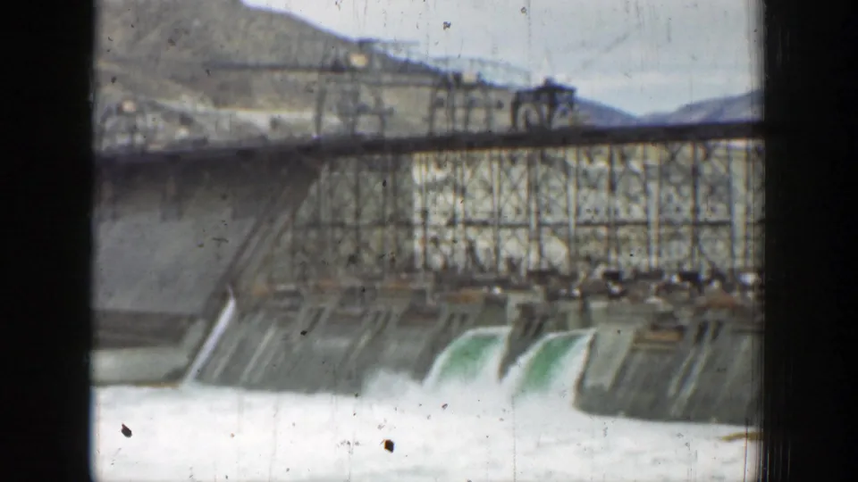 1937 California 8mm view of spillway water flowing down a concrete dam below a steel truss bridge and utility structures.