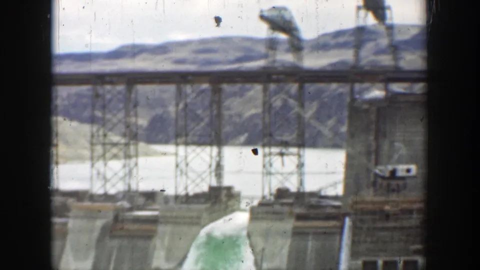 1937 California 8mm frame of bridge supports and dam towers over pale water, with hills in the background and visible film wear.