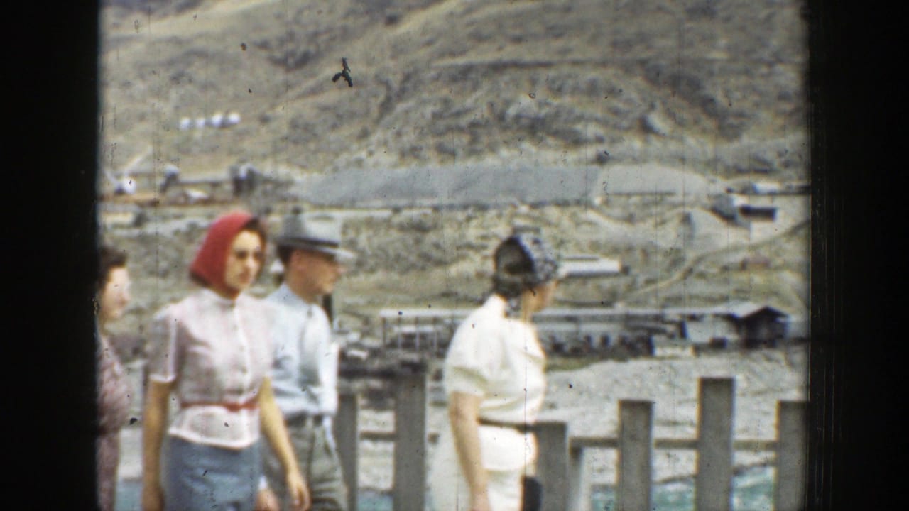 1937 California home-movie frame showing adults in period clothing walking past a grey fence against arid hills.
