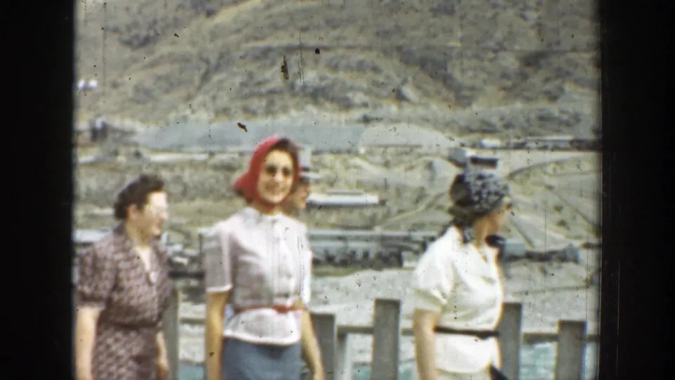 1937 California frame with women in dresses and a red headscarf walking along a grey fence in a dry landscape.
