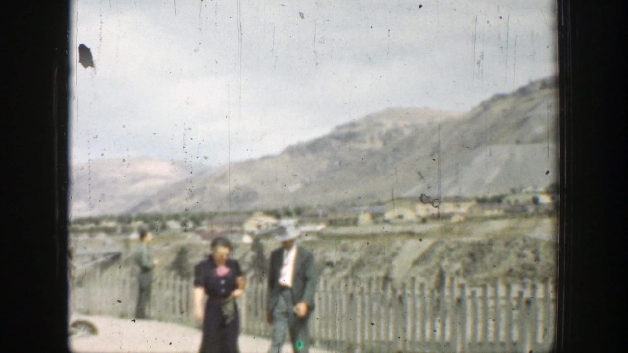 1937 California 8mm frame showing two adult tourists walking along a fenced dam overlook with hills and a distant settlement in view.