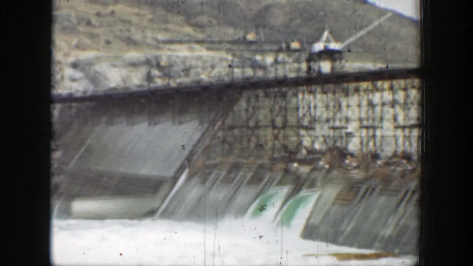 Wide 1937 California dam frame showing multiple spillway sections, metal framework, hillside terrain, and a small lookout structure near the crest.