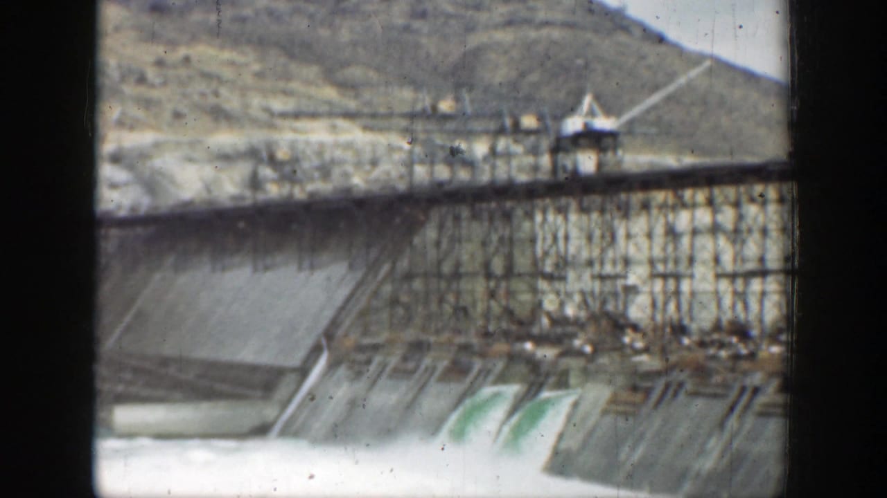 Concrete dam spillway with foaming water, scaffolding above, and hills in the background.