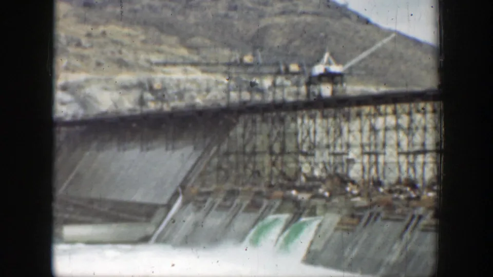 1937 California 8mm frame showing a dam or industrial spillway releasing foaming water below scaffolding and hillside terrain.