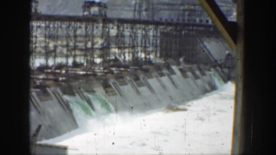 1937 California frame of a concrete dam face, crest scaffolding, and white water surging from the spillway into the river below.