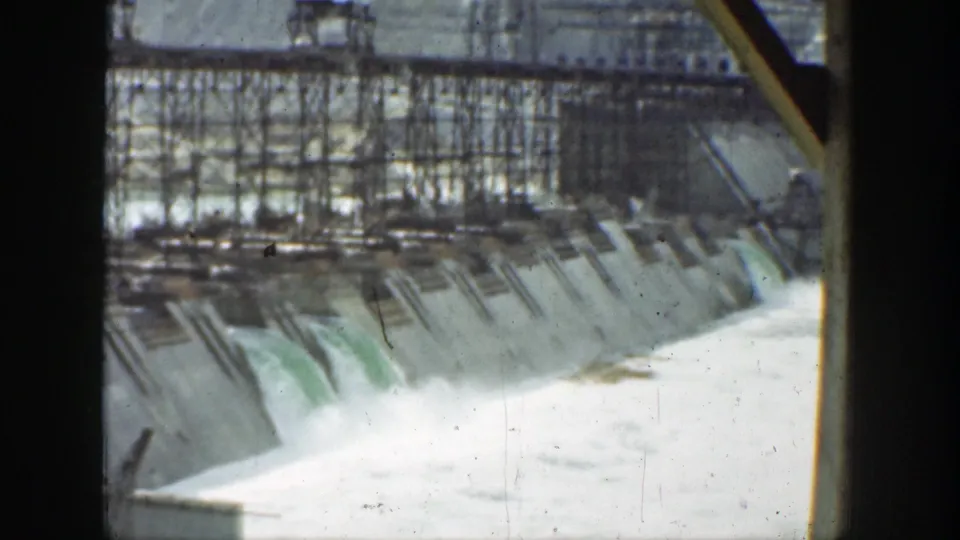 1937 California 8mm view across a dam spillway where foamy water rushes beneath dark scaffolding and concrete channels.