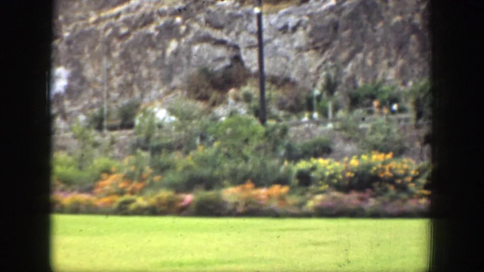 1937 California 8mm frame with bright lawn, colorful garden beds, shrubs, rocky hillside, and dark foreground framing.