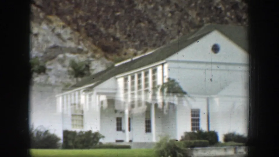 1937 California 8mm frame showing a white rural or facility building with tall windows, shrubs, steps, and a rocky slope behind it.