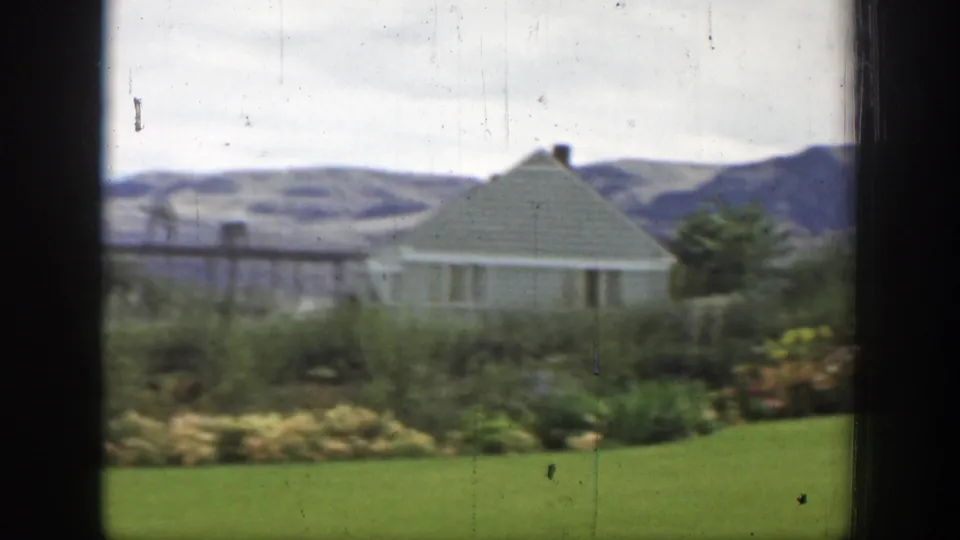 1937 California 8mm frame with a small white building, landscaped shrubs, green lawn, distant dam structures, and mountain ridges.