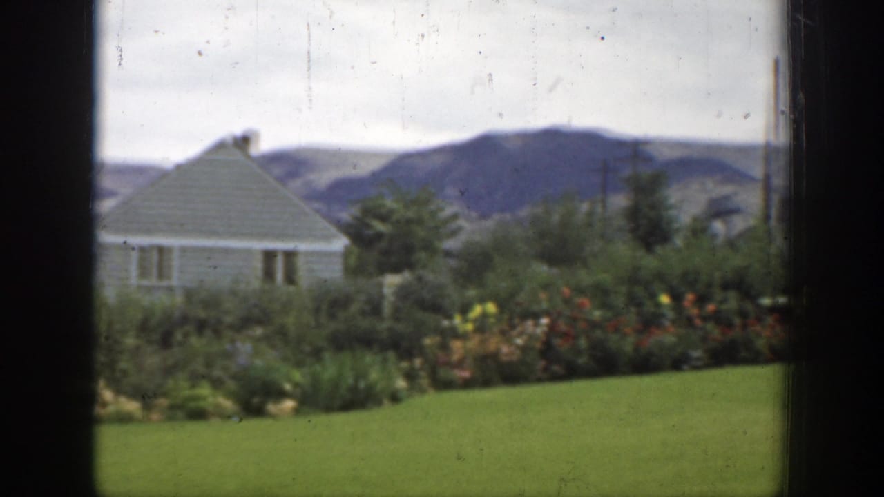 Green lawn and garden with a small house and hills beyond.