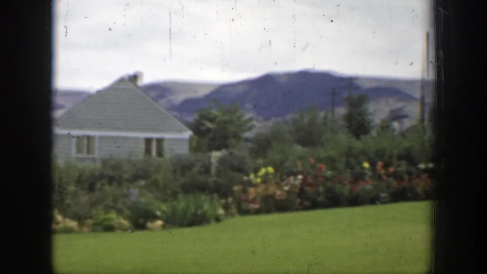 1937 California home-movie view of a green lawn, flower beds, a small house, utility poles, and distant hills.