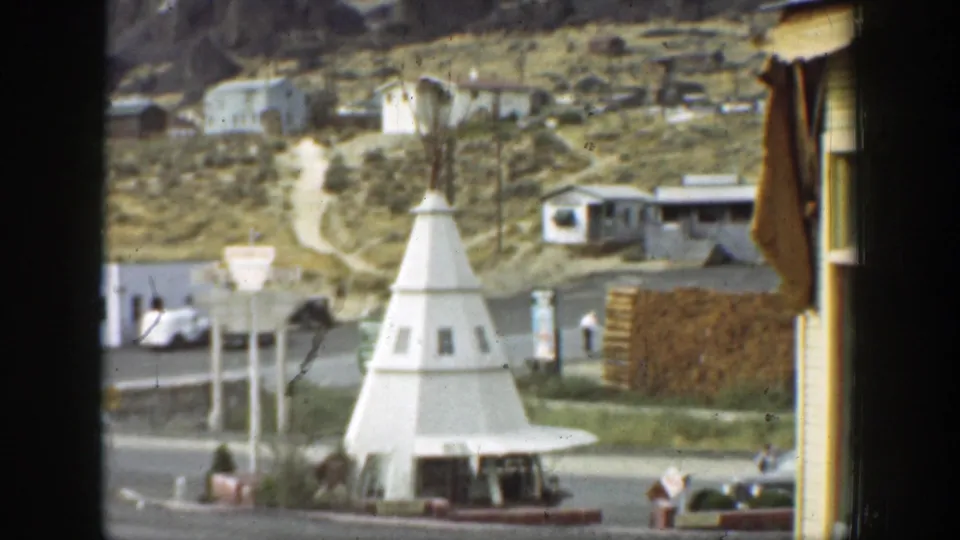 1937 California roadside view with a white cone-shaped structure, small buildings, cars, utility poles, and dry hills.