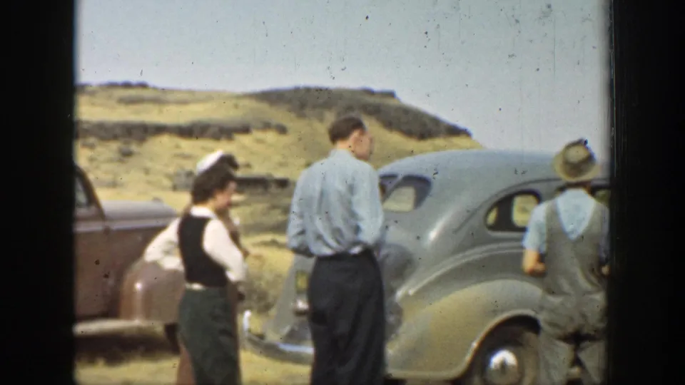 Adults in period clothing stand beside parked automobiles in a rural California hillside setting, with water visible beyond.