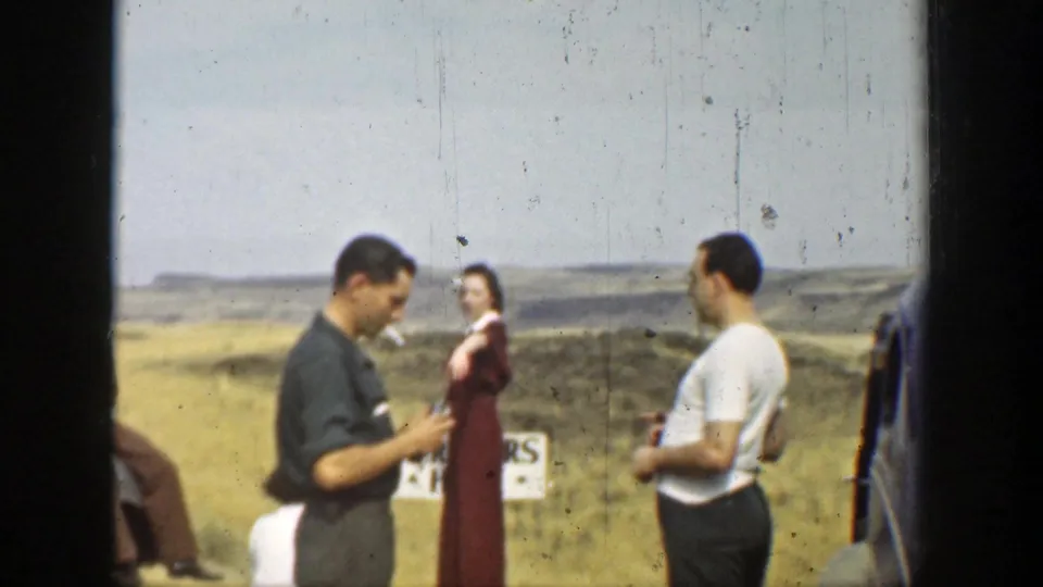 A 1937 home-movie frame shows two men near a parked car and a woman in a long dress in the open landscape.