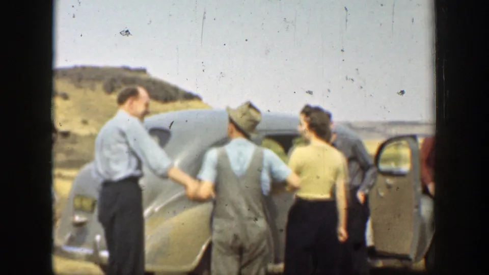 Adults in 1930s clothing cluster around a rounded automobile, including a man in overalls and a hat near an open door.