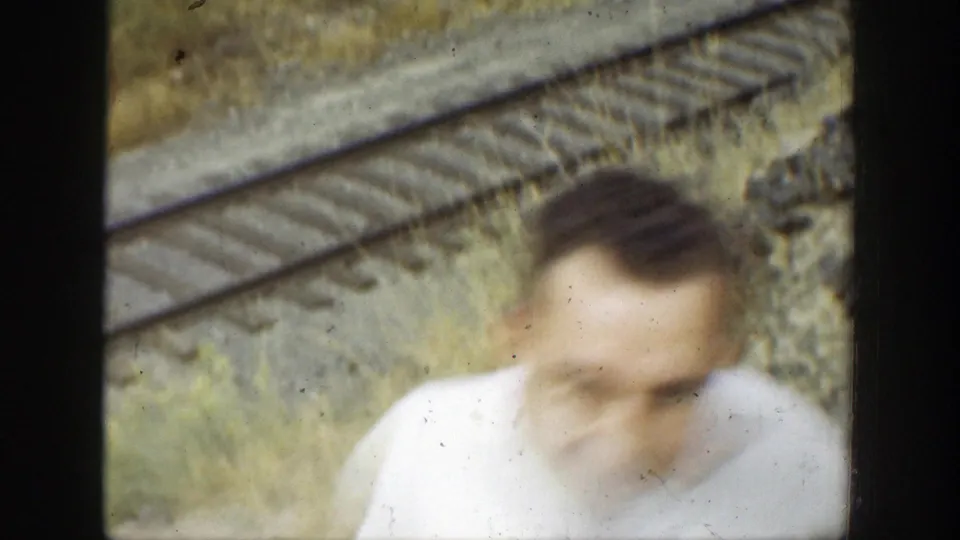 A blurred figure moves close to the camera, with railroad tracks and dry brush visible behind them.