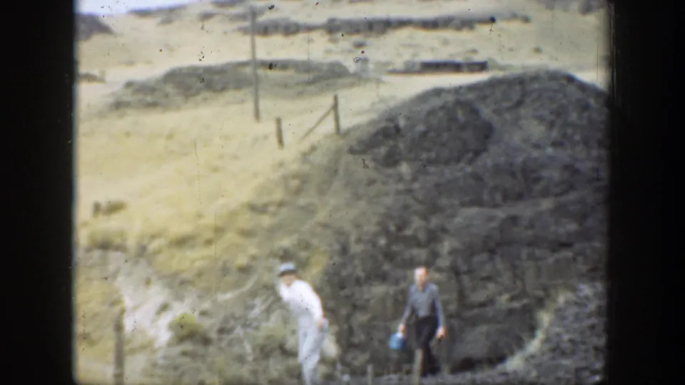 Two people appear near a dark rocky mound and sparse fence posts in dry California hillside terrain.