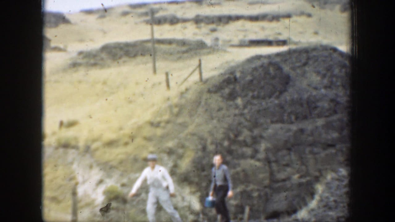Two men in period clothing walk across an arid California hillside near rocks and sparse fence posts.