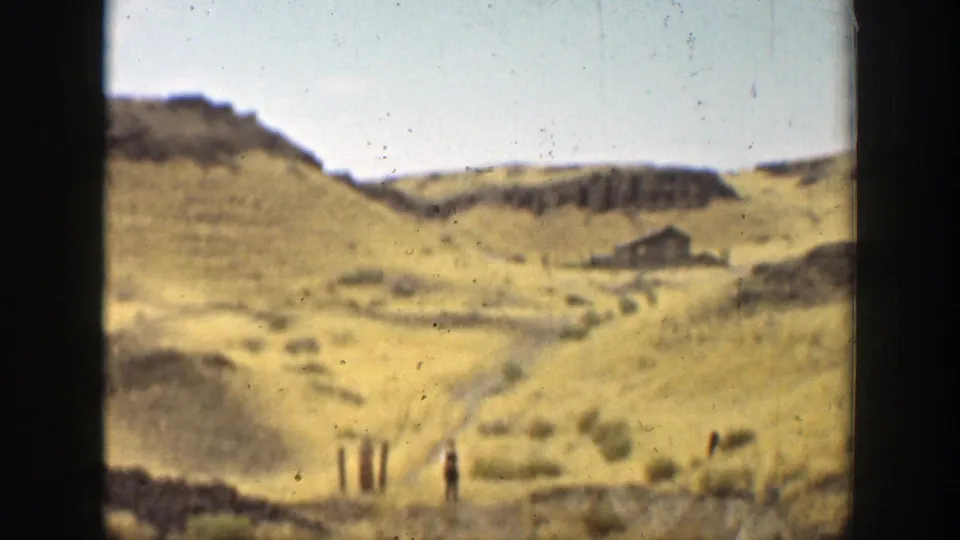Arid California hill landscape with scrub, fence posts, a valley path, and a small distant structure.