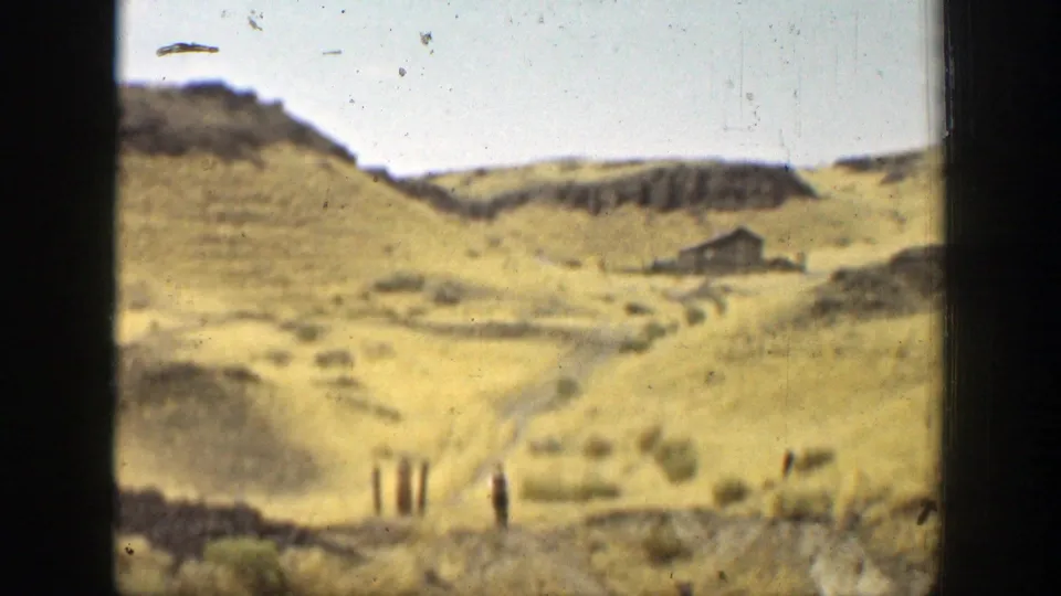 Wide view of dry California hills with scrub, fence posts, and a small cabin-like building in the valley.