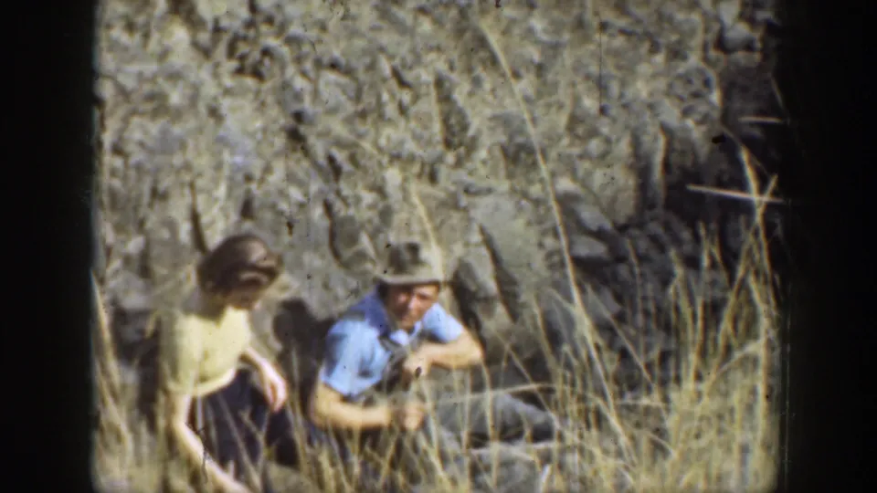 Two adults appear to crouch in dry grass near a rocky California canyon wall in this 1937 home-movie frame.