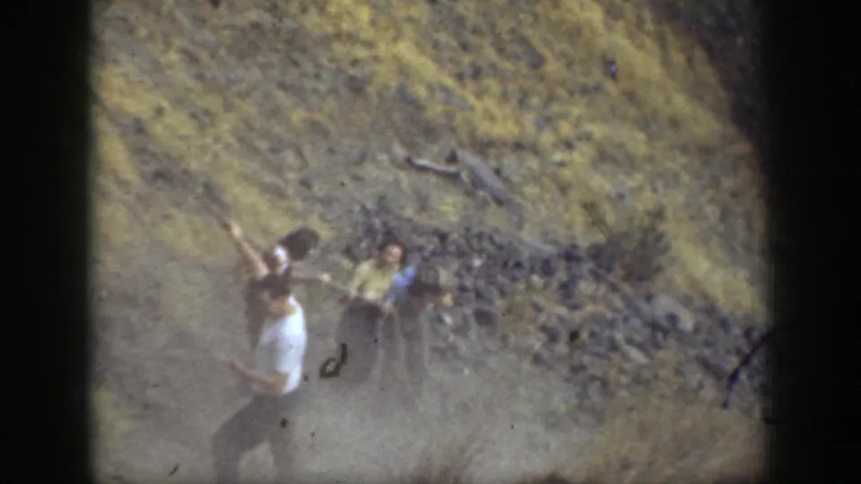 Home-movie frame of hikers on a rugged California trail, with one person using a rope or support on the slope.