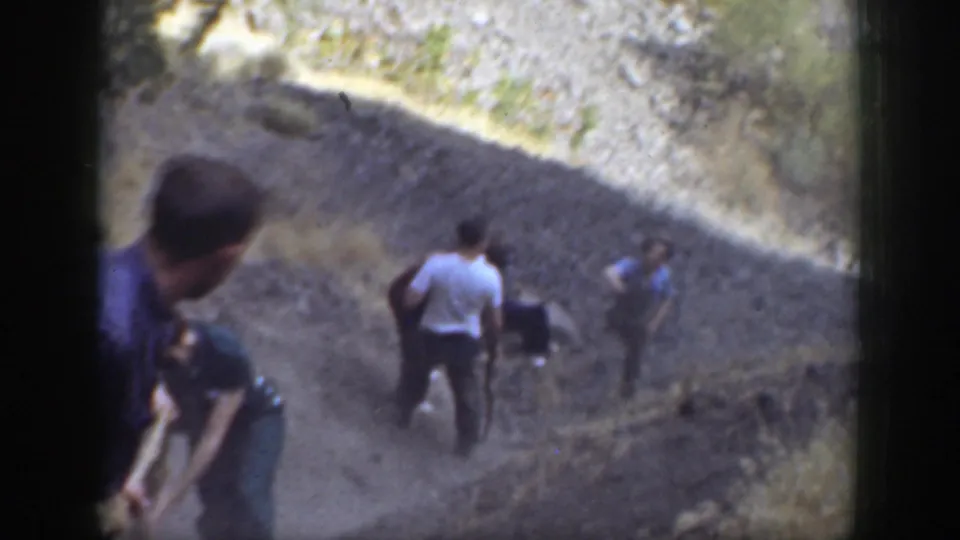 Home-movie frame from 1937 California showing companions gathered on a dusty rocky trail after an apparent fall.