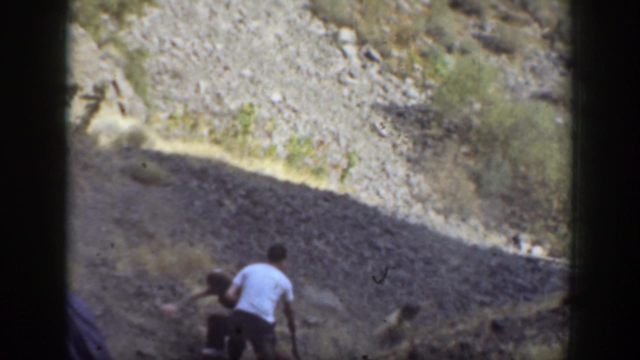 1937 California home-movie frame showing adults on a dry rocky slope as a man in a light shirt assists a woman climbing upward.