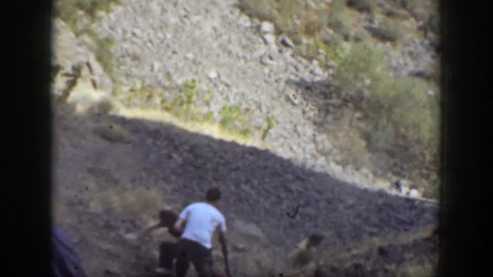 1937 California home-movie frame showing adults on a dry rocky slope as a man in a light shirt assists a woman climbing upward.