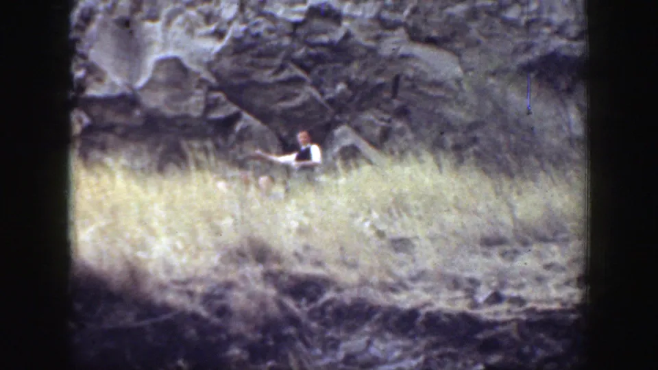 1937 California home-movie frame with a distant adult near dry grass and exposed rock on a sunlit hillside.
