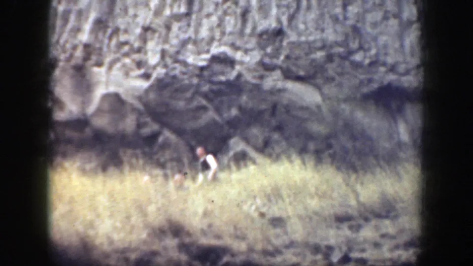 1937 California home-movie frame showing a distant adult partly obscured by dry grass below a rugged rock face.