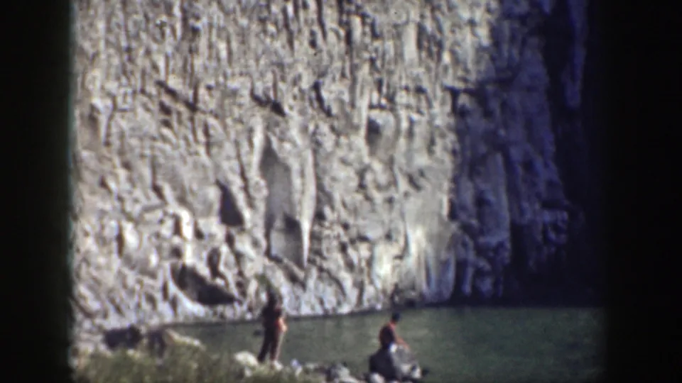 Two figures appear near a small body of water beneath a vertical rock wall in 1937 California home-movie footage.