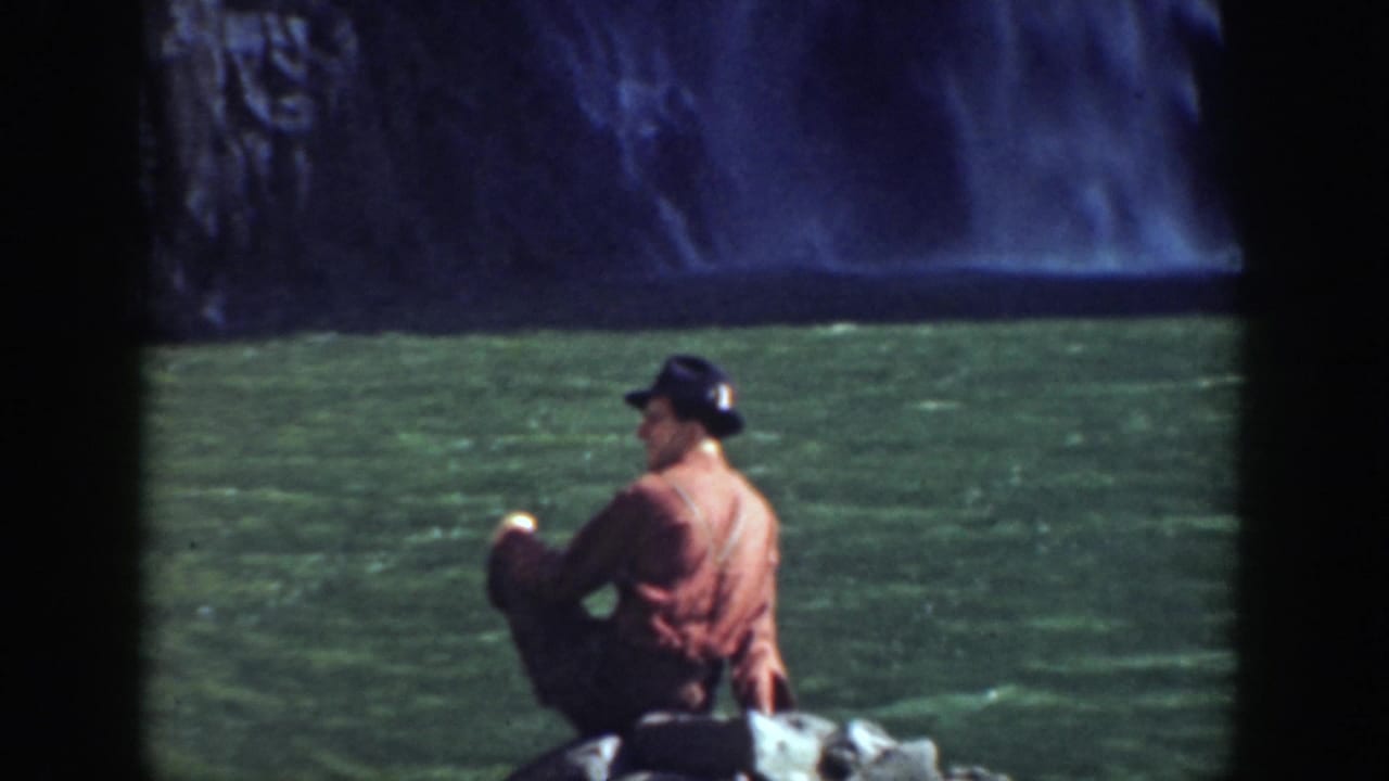 Man in a dark cap and reddish shirt sitting on rocks beside green water and a waterfall.