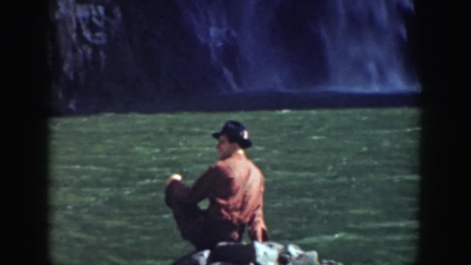 1937 California 8mm frame of a seated man in a cap looking toward a waterfall and green pool from a rocky shore.