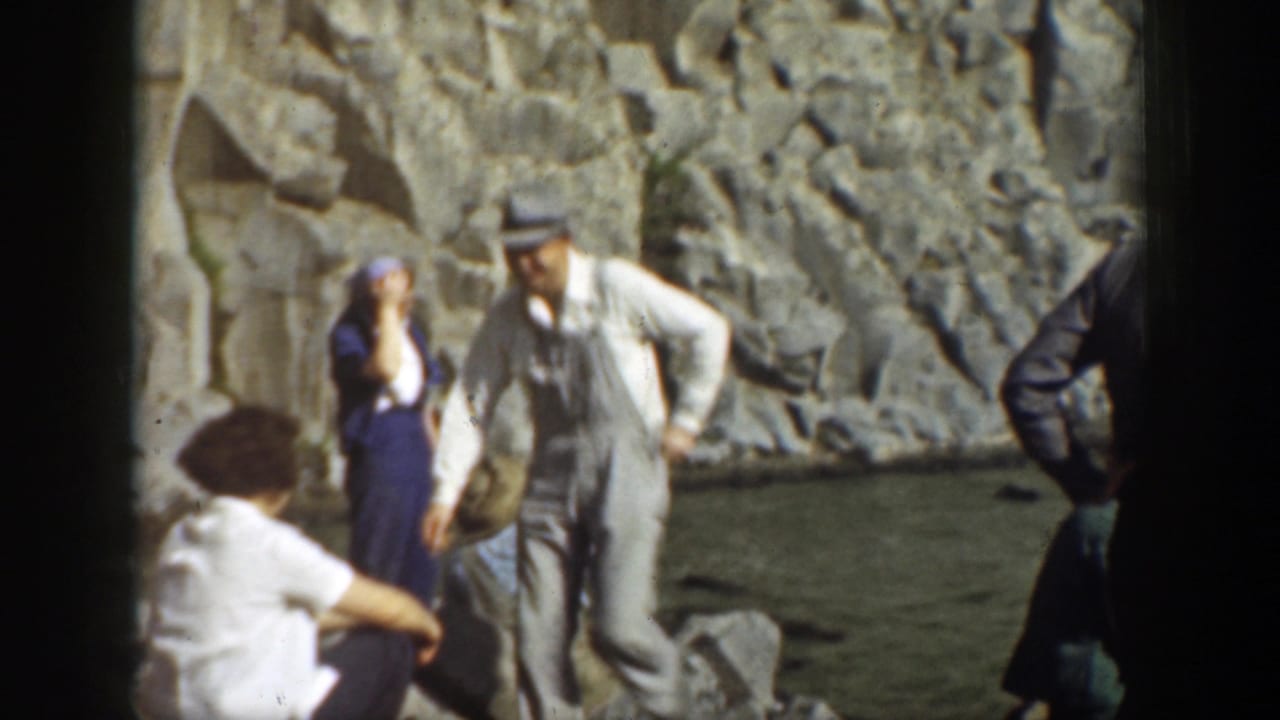 Adults in period clothing standing and sitting near rocks beside the waterfall pool.