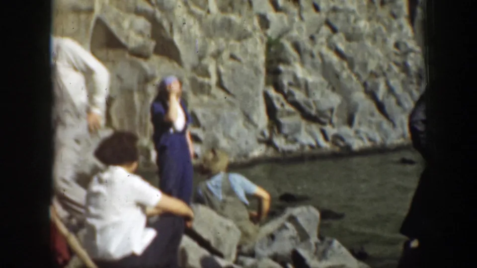 A woman in a dark dress stands among adults on the rocky shoreline in a 1937 California waterfall home movie frame.