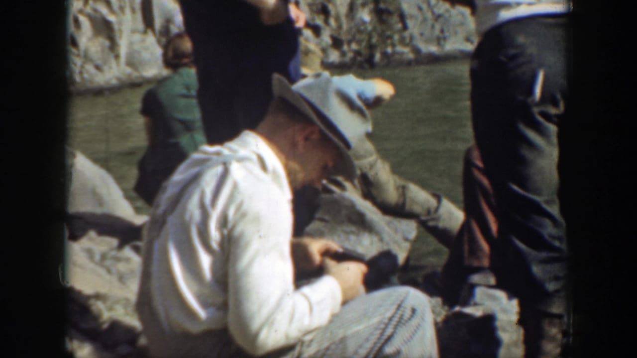 Man in a fedora sits on rocks near water while other adults stand nearby.