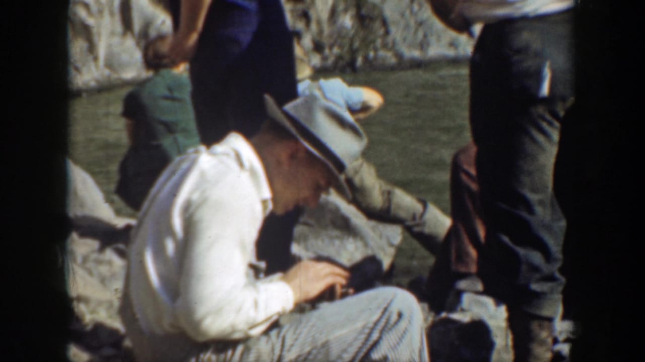 Man in a fedora looks down while seated on rocks beside standing visitors.